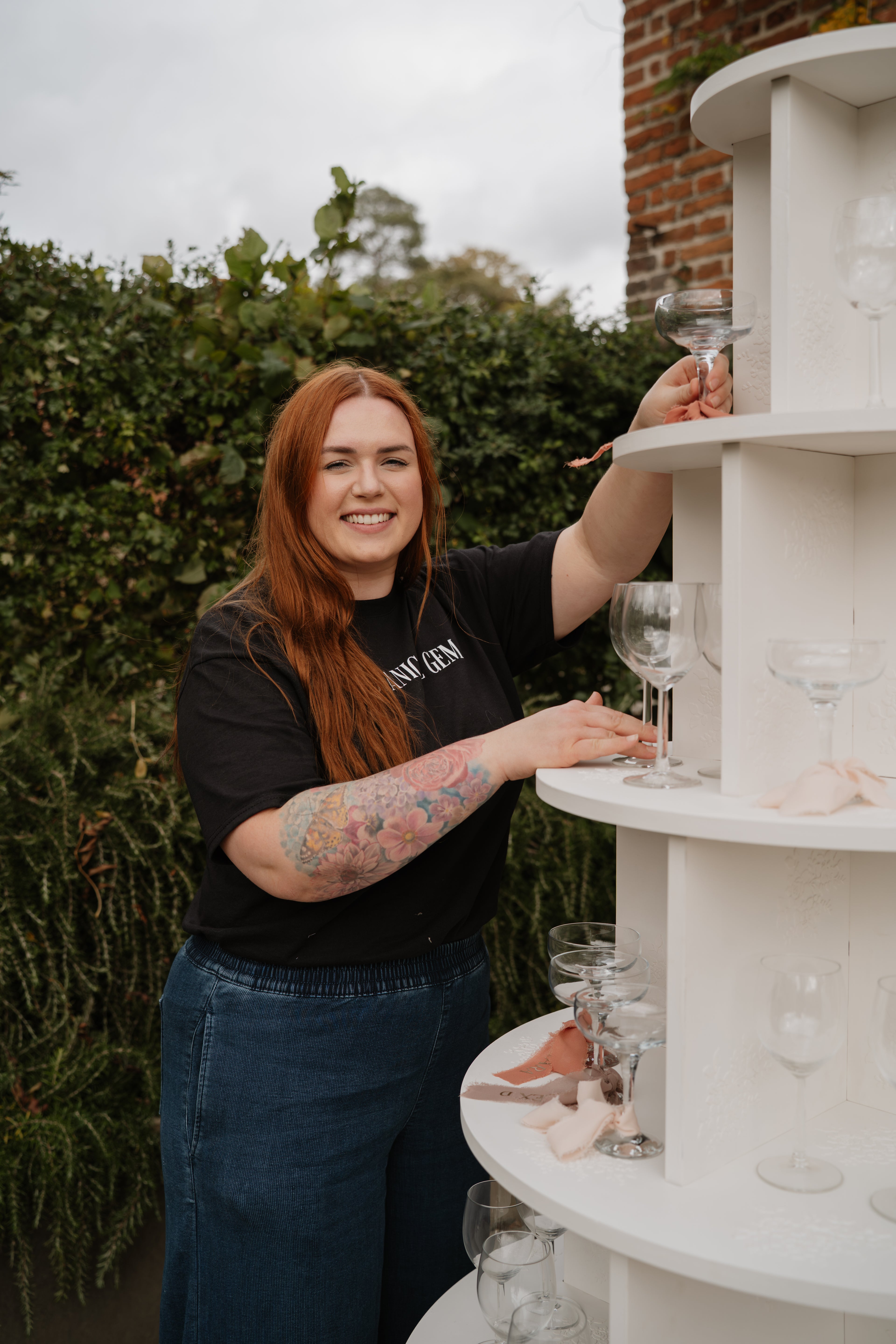 Lady with ginger hair and a friendly smile is arranging wine glasses on a large white display unit outdoors